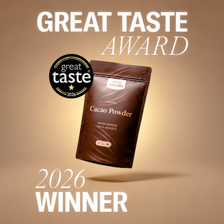 A brown stand-up pouch of cacao powder floating on a warm beige gradient background, with large white text reading 'GREAT TASTE AWARD' at the top and '2026 WINNER' at the bottom. A circular 'great taste 2026' badge appears to the left of the pouch.
